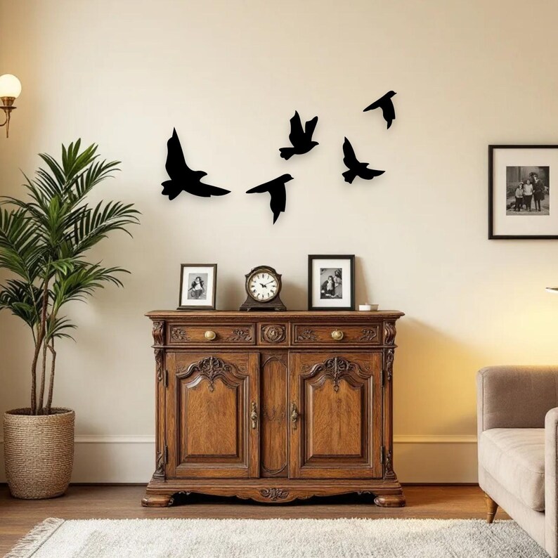 May include: A wooden cabinet with ornate carvings, topped with a clock and framed photos. Above, black bird silhouettes are arranged on a cream-colored wall. A potted plant and a framed picture are also visible.