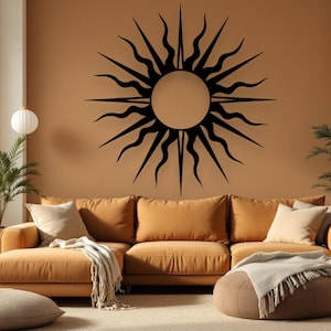 May include: A large, black sunburst wall art decorates a tan wall above a mustard-coloured sectional sofa. The sun design features a central circle with radiating, stylized flames. The room is decorated with neutral-toned pillows and a blanket.