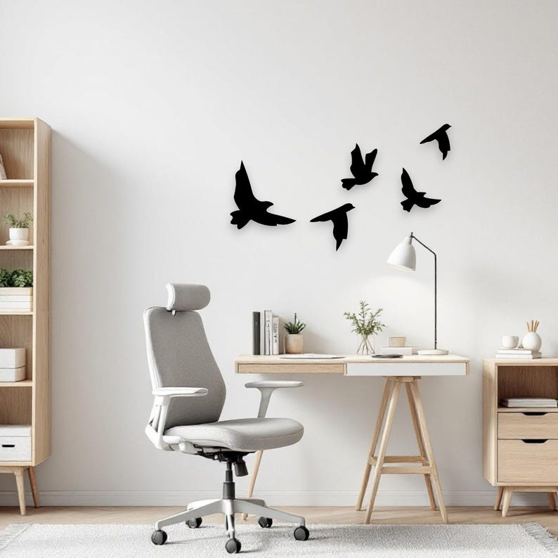 May include: Black silhouette birds in flight wall decor. The birds are mounted on a white wall above a desk and office chair. The desk is made of light wood and has a white top. A lamp and small plant are on the desk.