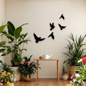 May include: Black metal bird wall art in various flying poses. The birds are mounted on a white wall, creating a minimalist and modern decorative accent. The scene includes potted plants and a wooden table.