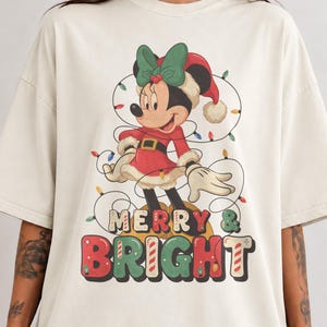 以下が含まれることがあります： オフホワイトのオーバーサイズTシャツ。サンタ帽と緑のリボンを身につけ、カラフルなクリスマスライトに包まれたミニーマウスのグラフィックが描かれています。キャラクターの下には、大きくスタイリッシュな多色の文字で「MERRY & BRIGHT」と記されています。