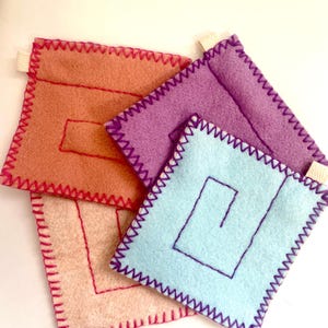May include: Three square felt pouches in peach, purple, and light blue. Each pouch features a small white fabric tab on the side and is stitched with a contrasting zigzag border. A simple rectangular or spiral line pattern is embroidered on the front.