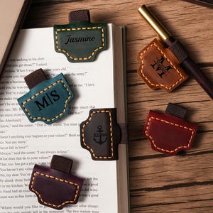 May include: A collection of leather bookmarks in various colors, including green, teal, brown, and burgundy. Each bookmark features a unique design and personalized text, with a pen resting on top of the book. The bookmarks are stitched with contrasting thread.