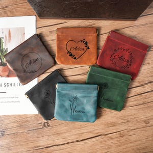 May include: A collection of small, leather coin purses in various colors, including brown, teal, and green. Each purse features a unique design, such as a floral pattern or a name, and a metal clasp. The purses are arranged on a wooden surface.