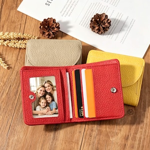 Puede incluir: Una cartera plegable de cuero rojo texturizado con una ventana transparente que muestra una foto de tres personas, múltiples ranuras para tarjetas y un cierre de broche metálico. Dos carteras adicionales en beige y amarillo están detrás.