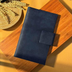 May include: A navy blue leather journal with a rectangular flap closure. The journal is placed on a wooden surface with a straw hat and wheat stalks in the background. The journal is a dark blue color.