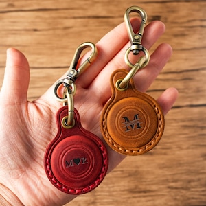 Puede incluir: Dos llaveros de cuero con cierres metálicos. Uno es rojo con las iniciales "M♡R", el otro es marrón con el nombre "Madison" y la letra "M" grabada. Ambos tienen un diseño circular y se sostienen en una mano.