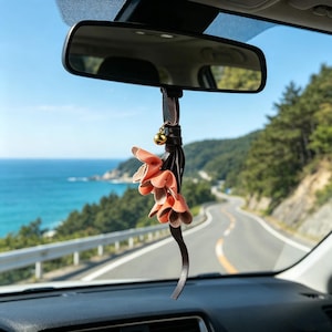 May include: A car rearview mirror hanging ornament featuring a dark brown leather strap, a small gold-toned bell, and a cluster of layered, peach-colored leather flower petals that dangle vertically from the mirror mount.