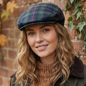 Puede incluir: Gorra plana a cuadros azul marino, verde y rojo. La persona lleva un jersey marrón y una chaqueta verde oscuro. El fondo es una pared de ladrillo con hiedra.
