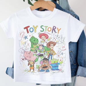 May include: White t-shirt featuring a colorful sketch of Toy Story characters, including Woody, Buzz Lightyear, and Rex. The words "TOY STORY" are written across the top in various colors. The shirt is hanging on a wooden hanger.