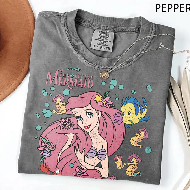 Puede incluir: Una camiseta de manga corta gris carb&oacute;n con un estampado gr&aacute;fico de Ariel de La Sirenita con el pelo rosa, rodeada de burbujas, flores y criaturas marinas. La camiseta tiene cuello redondo y una etiqueta de la marca Comfort Colors.