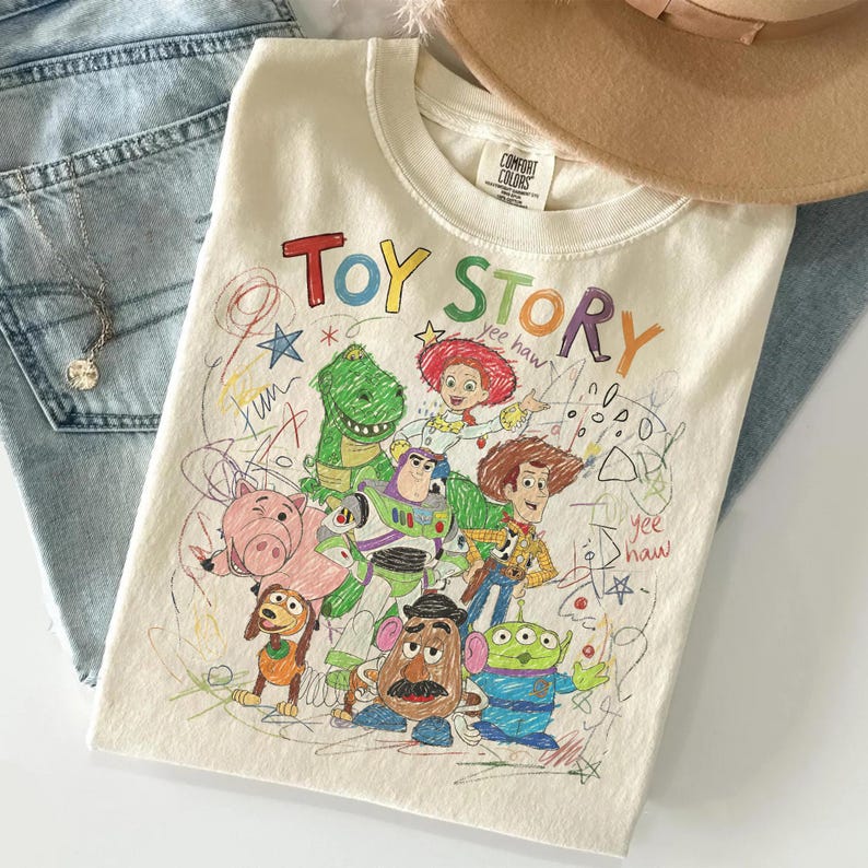 May include: Cream-colored t-shirt featuring a colorful graphic of Toy Story characters, including Woody, Buzz Lightyear, and Rex. The words "Toy Story" are written in a playful, multi-colored font. The shirt is folded on top of blue jeans.