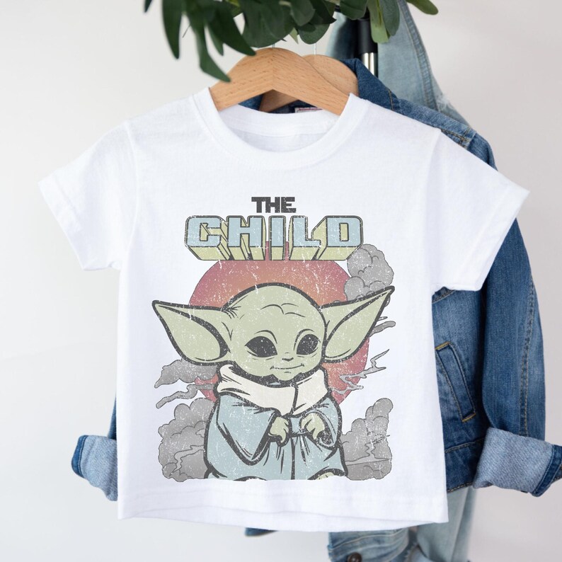 May include: A white short-sleeve t-shirt featuring a distressed graphic of a green creature with large ears and a tan robe. Above the character, the text reads THE CHILD in block letters. The background includes a red sun and gray clouds.