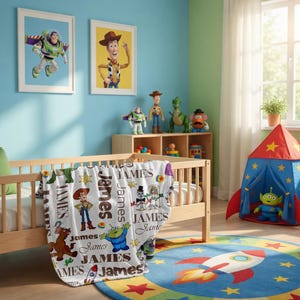 Puede incluir: Una manta de forro polar blanca con un patr&oacute;n repetitivo de personajes de Toy Story, incluyendo a Woody, Buzz Lightyear y un alien&iacute;gena, intercalados con el nombre James en varias fuentes y colores, sobre una cuna de madera clara.