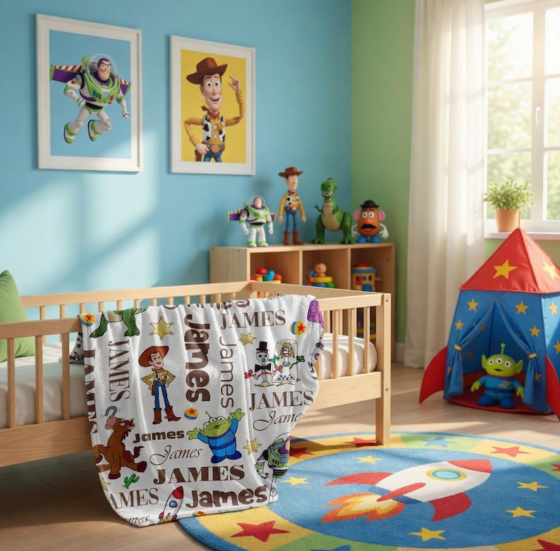 Puede incluir: Una manta de forro polar blanca con un patr&oacute;n repetitivo de personajes de Toy Story, incluyendo a Woody, Buzz Lightyear y un alien&iacute;gena, intercalados con el nombre James en varias fuentes y colores, sobre una cuna de madera clara.