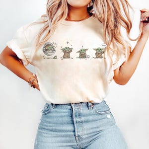 May include: Cream-colored t-shirt featuring a cartoon sequence of a character from a popular science fiction series. The character is depicted in various poses, with the word "Cool" in a speech bubble.