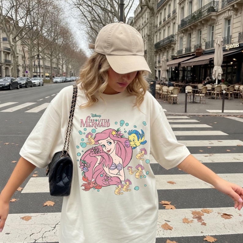 Puede incluir: Una gorra de b&eacute;isbol beige y una camiseta color crema con un estampado gr&aacute;fico de Ariel de La Sirenita con cabello rosa, un top de concha morado y personajes de peces y caballitos de mar amarillos, junto a un bolso de hombro negro acolchado.