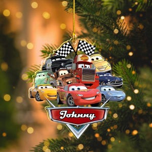 May include: A Cars-themed Christmas ornament featuring various animated car characters, including Lightning McQueen. The ornament has a gold string for hanging and a banner that reads "Johnny". Checkered flags are at the top.