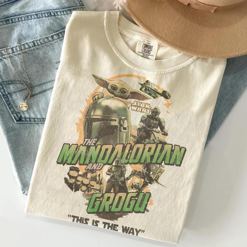 May include: Cream-colored t-shirt featuring a graphic of The Mandalorian and Grogu from Star Wars. The design includes the text "THE MANDALORIAN AND GROGU" and the phrase "THIS IS THE WAY". A tan hat and blue jeans are also visible.