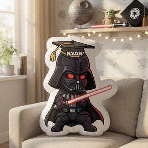 May include: A plush pillow shaped like Darth Vader, wearing a graduation cap with the name "RYAN". The character is holding a red lightsaber. The pillow is on a beige couch in a room with Star Wars-themed decor.