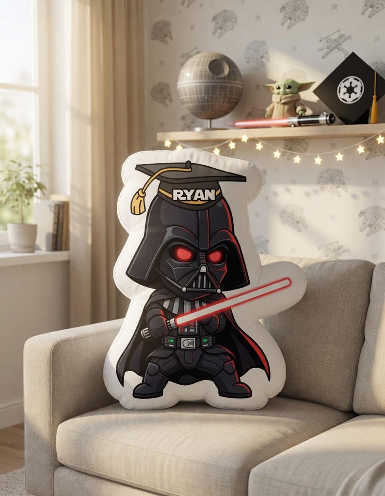 May include: A plush pillow shaped like Darth Vader, wearing a graduation cap with the name "RYAN". The character is holding a red lightsaber. The pillow is on a beige couch in a room with Star Wars-themed decor.