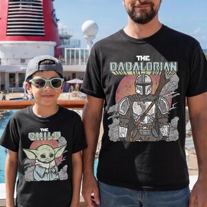 May include: Two black t-shirts featuring graphic prints. The larger shirt displays a Mandalorian character with the text THE DADALORIAN. The smaller shirt features a small green creature with large ears and the text THE CHILD, both set against a red circle.