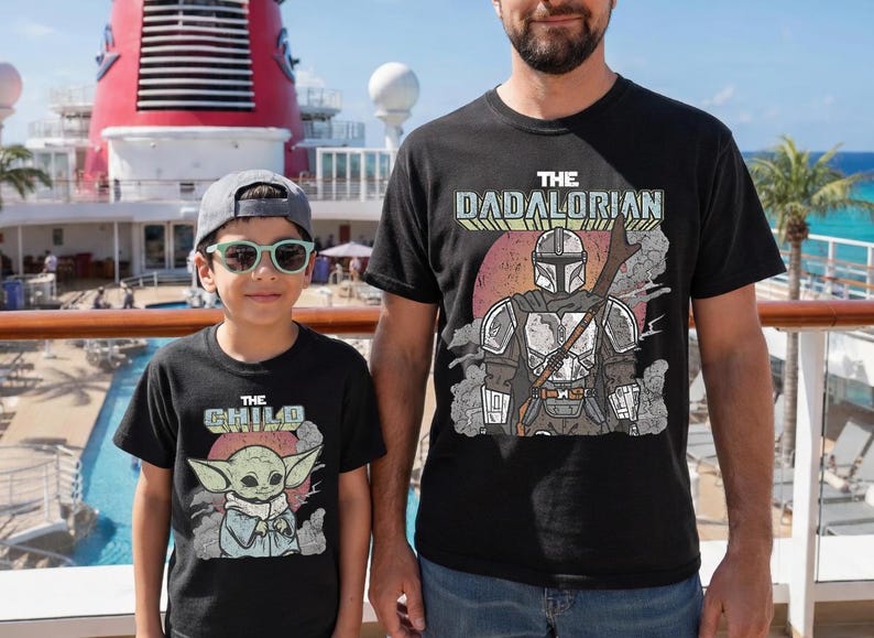 May include: Two black t-shirts featuring graphic prints. The larger shirt displays a Mandalorian character with the text THE DADALORIAN. The smaller shirt features a small green creature with large ears and the text THE CHILD, both set against a red circle.