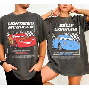 May include: Two charcoal gray t-shirts featuring characters from the movie Cars. One shirt displays Lightning McQueen, a red race car, with text. The other shirt shows Sally Carrera, a blue car, with text. Both shirts have a distressed look.