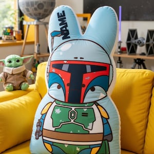 May include: A light blue bunny-shaped cushion with a cartoon illustration of Boba Fett's helmet and armour. The cushion has the word "NAME" printed on one ear. The cushion is on a yellow sofa.