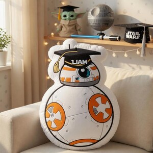 May include: A white BB-8 plush pillow with orange and black details, wearing a graduation cap with the name "LIAM". The pillow is on a beige couch, with Star Wars themed decor on a shelf in the background.