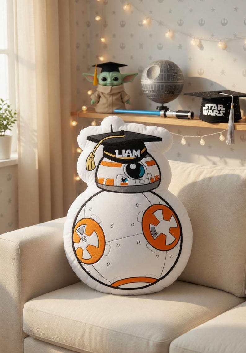 May include: A white BB-8 plush pillow with orange and black details, wearing a graduation cap with the name "LIAM". The pillow is on a beige couch, with Star Wars themed decor on a shelf in the background.