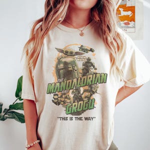 May include: Cream-colored t-shirt featuring a graphic design from the Star Wars series, "The Mandalorian." The design includes images of Grogu, the Mandalorian, and other characters, with the text "The Mandalorian and Grogu" and "This is the Way."