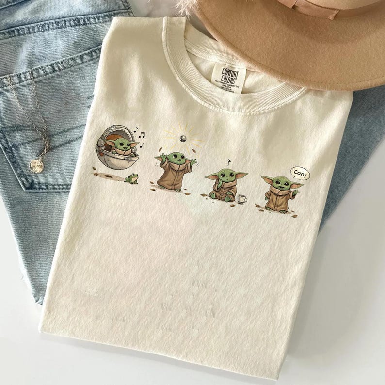 May include: Cream-colored Comfort Colors t-shirt featuring a graphic of a character from a popular science fiction series. The graphic shows the character in various poses. A tan hat and a pair of blue jeans are in the background.