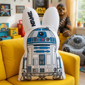 May include: A white and blue R2-D2 shaped cushion with bunny ears, on a yellow sofa. The cushion has the text "NAME" in black. The design is inspired by the Star Wars character R2-D2.