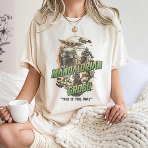 May include: Cream-colored t-shirt featuring a graphic design from the Star Wars series, "The Mandalorian and Grogu." The design includes images of characters and the text "THIS IS THE WAY" in green and white. The shirt is a casual wear item.