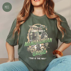 May include: Moss green t-shirt featuring a graphic from the Star Wars series, "The Mandalorian and Grogu." The design includes the Mandalorian's helmet, Grogu, and the text "THIS IS THE WAY." The shirt is a relaxed fit.