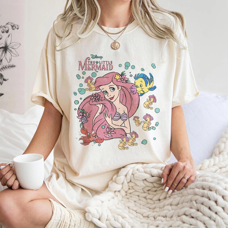 Puede incluir: Una camiseta de color crema con un estampado gr&aacute;fico de Ariel de La Sirenita con el pelo rosa, rodeada de Flounder, Sebasti&aacute;n y varios caballitos de mar entre burbujas y flores de color verde azulado. El texto dice Disney The Little Mermaid.
