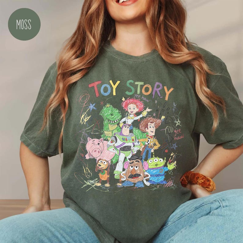 May include: Moss green t-shirt featuring a colorful graphic of Toy Story characters. The design includes the words "Toy Story" in a playful font, along with drawings of Woody, Buzz Lightyear, and other characters. The shirt is a comfortable, casual style.