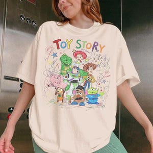 May include: Cream-colored t-shirt featuring a colorful, hand-drawn graphic of characters from the movie "Toy Story." The text "Toy Story" is at the top. The design includes Woody, Buzz Lightyear, and other characters.