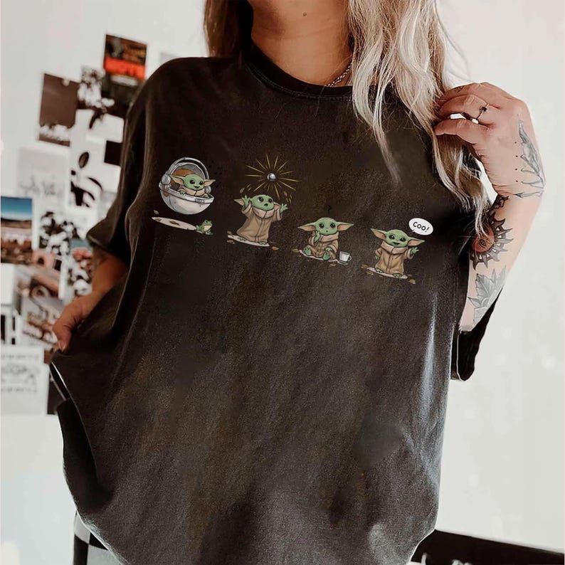 May include: Dark gray t-shirt featuring a graphic of a character from a popular science fiction series. The graphic shows the character in four different poses. The last pose has the word "Cool!" in a speech bubble. The t-shirt is a crew neck.