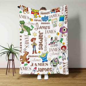Puede incluir: Una manta de forro polar blanca con un patr&oacute;n repetitivo de personajes de Toy Story, incluyendo a Woody, Buzz Lightyear, Rex y Jessie, intercalados con el nombre James en texto marr&oacute;n y el logotipo de Toy Story 5 en azul, amarillo y rojo.