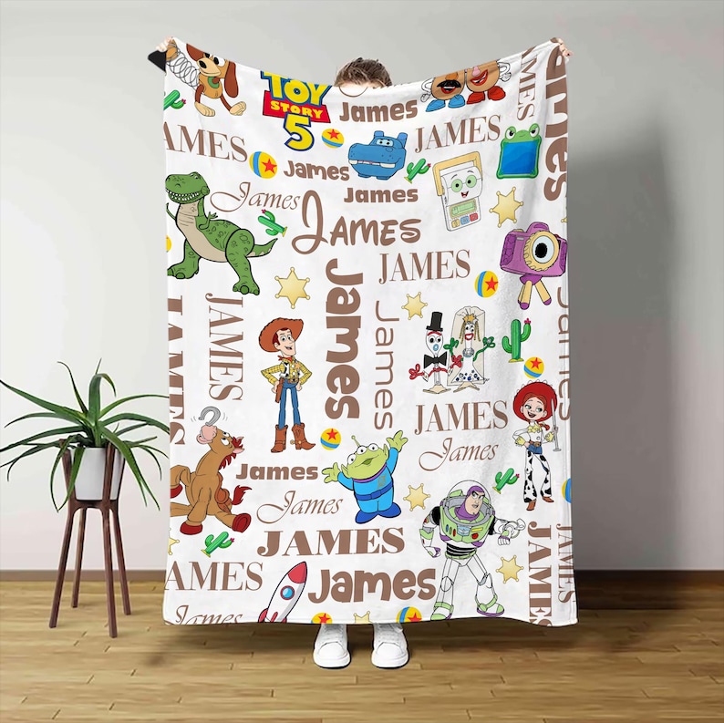 Puede incluir: Una manta de forro polar blanca con un patr&oacute;n repetitivo de personajes de Toy Story, incluyendo a Woody, Buzz Lightyear, Rex y Jessie, intercalados con el nombre James en texto marr&oacute;n y el logotipo de Toy Story 5 en azul, amarillo y rojo.
