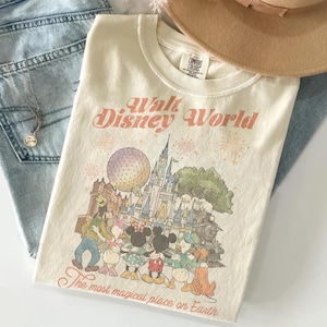 Puede incluir: Camiseta color crema con un gr&aacute;fico de personajes de Disney World y el texto "Walt Disney World" y "The most magical place on Earth." La camiseta est&aacute; doblada sobre unos vaqueros azules, con un collar y un sombrero beige.