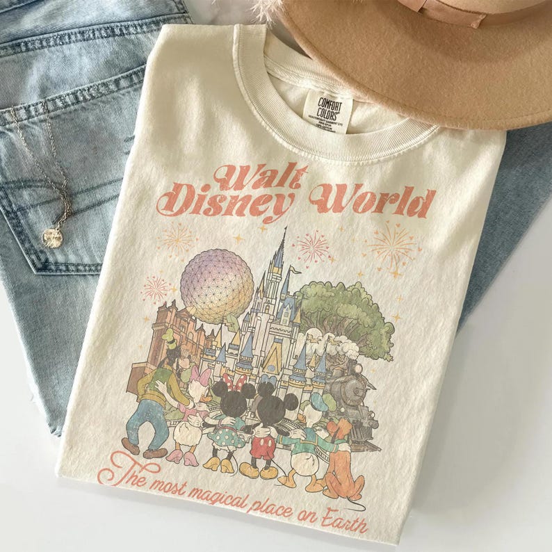Puede incluir: Camiseta color crema con un gr&aacute;fico de personajes de Disney World y el texto "Walt Disney World" y "The most magical place on Earth." La camiseta est&aacute; doblada sobre unos vaqueros azules, con un collar y un sombrero beige.
