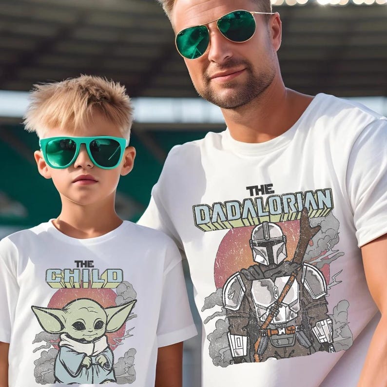 May include: Two white t-shirts featuring graphic prints. The larger shirt displays a Mandalorian character with the text THE DADALORIAN. The smaller shirt shows a green creature with large ears and the text THE CHILD. Both shirts have a retro design.