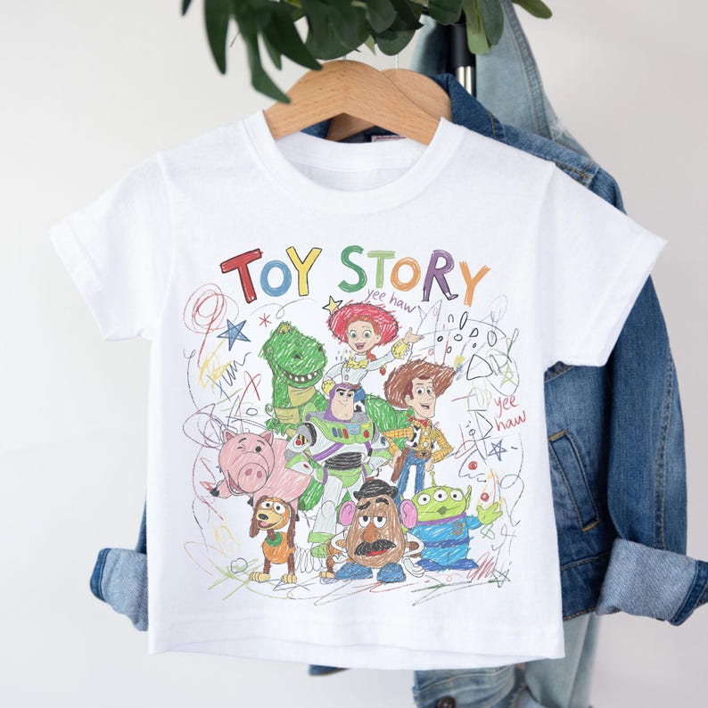 May include: White t-shirt featuring a colorful sketch of Toy Story characters, including Woody, Buzz Lightyear, and Rex. The words "TOY STORY" are written across the top in various colors. The shirt is hanging on a wooden hanger.