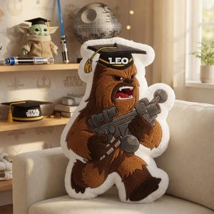 May include: A plush Chewbacca pillow wearing a graduation cap with the name "LEO" on it. The brown and white pillow is holding a blaster and is sitting on a beige couch. The background includes Star Wars themed decor.