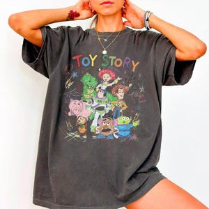 May include: Dark gray t-shirt featuring a colorful graphic of characters from the animated film "Toy Story." The design includes Woody, Buzz Lightyear, and other characters, with the words "Toy Story" in a playful font.