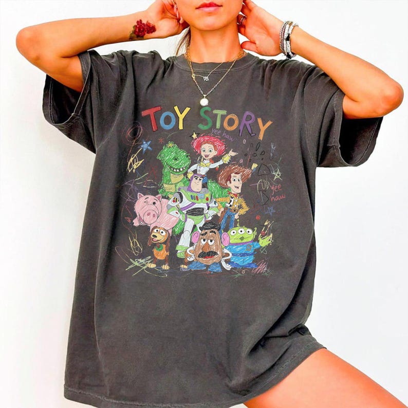 May include: Dark gray t-shirt featuring a colorful graphic of characters from the animated film "Toy Story." The design includes Woody, Buzz Lightyear, and other characters, with the words "Toy Story" in a playful font.