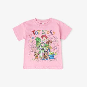 May include: Pink short-sleeved t-shirt featuring the text "TOY STORY" above a colorful illustration of characters from the animated movie. The design includes Woody, Buzz Lightyear, and other characters. The shirt is made of a soft material.